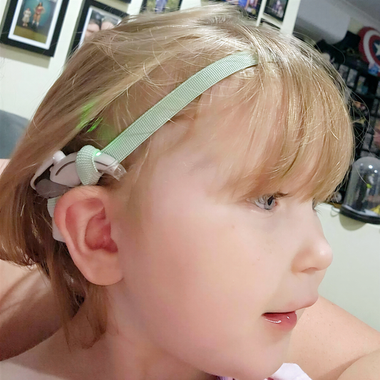 A young child with blonde hair wearing a green cochlear implant hearing aid headband and smiling.