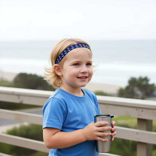 A young blonde girl wearing a blue t-shirt and a navy blue anchor-patterned headband for a cochlear implant or hearing aid, holding a stainless steel travel tumbler on a wooden boardwalk with a blurred beach background.