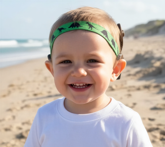A smiling toddler wearing a green dinosaur-print cochlear implant hearing aid headband, standing on a sandy beach with the ocean in the background.