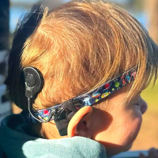 A young child wearing a colorful headband with a hearing aid attached, smiling and looking at the camera.