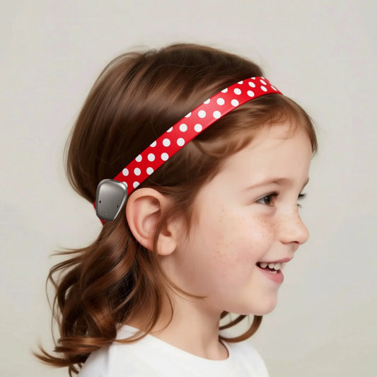 A red ponto baha hearing aid headband with white polka dots and a dark gray clip, shown against a white background with a faint gray silhouette of a face.