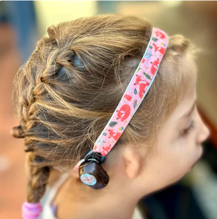 Child with light brown hair wearing a hearing aid headband with a peach and orange fox print on it. Side profile.