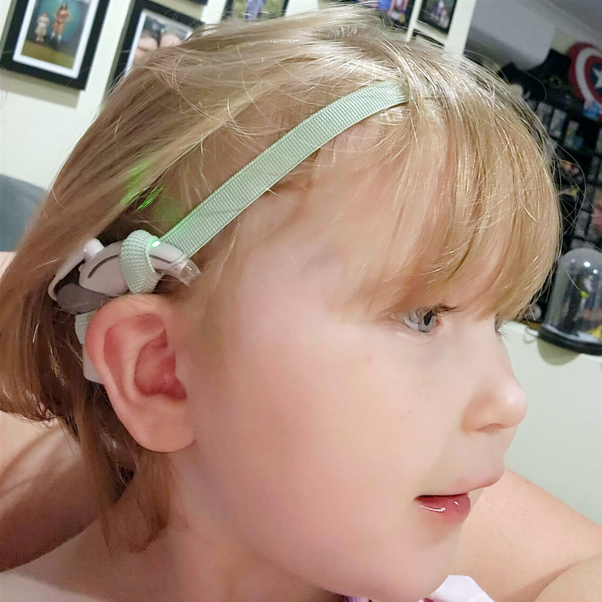 A young child with blonde hair wearing a green cochlear implant hearing aid headband and smiling.