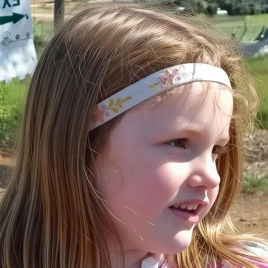 A young girl with long light brown hair wearing a white fabric hearing aid headband decorated with a yellow and pink floral pattern, photographed outdoors in bright sunlight.