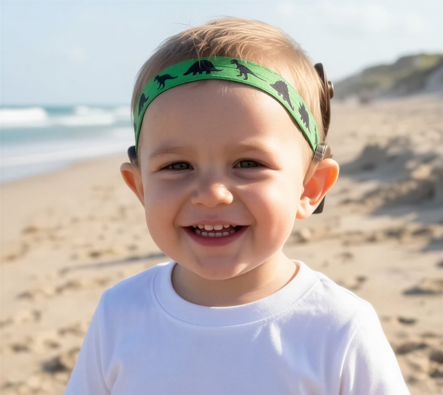A smiling toddler wearing a green dinosaur-print cochlear implant hearing aid headband, standing on a sandy beach with the ocean in the background.