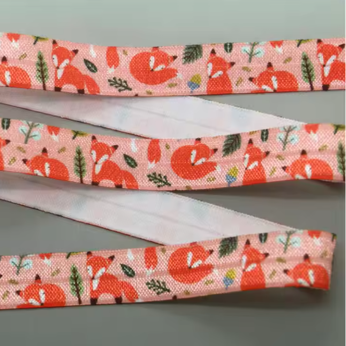 A close-up of a patterned elastic strap or band laid diagonally on a gray background. The fabric features a repeating print of orange foxes, green leaves, and small floral motifs on a peach colour base.