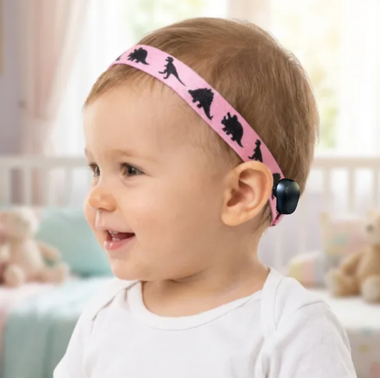 A baby wearing a pink Ponto BAHA softband headband with black dinosaur silhouettes, photographed in a softly lit nursery setting