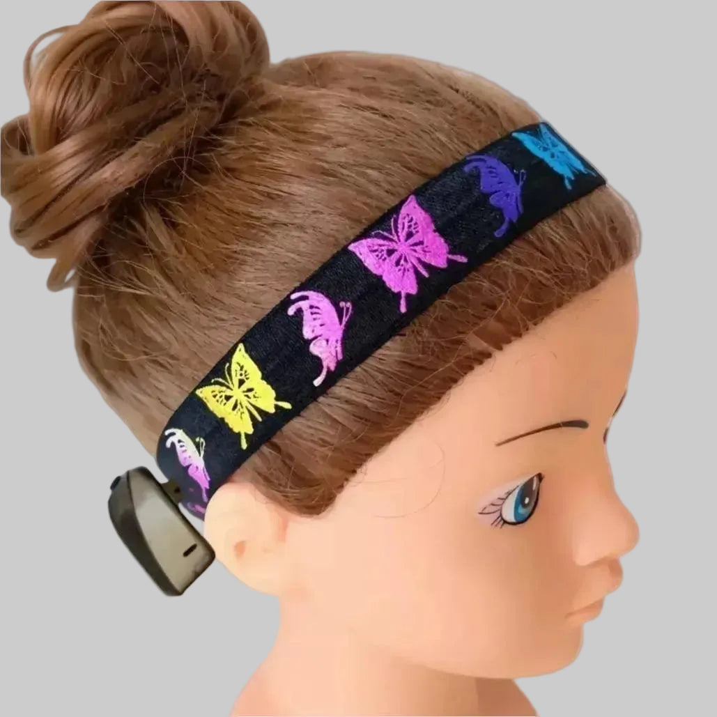 A black fabric cochlear implant headband with colourful butterfly motifs, shown worn on a doll mannequin head against a solid light pink background.