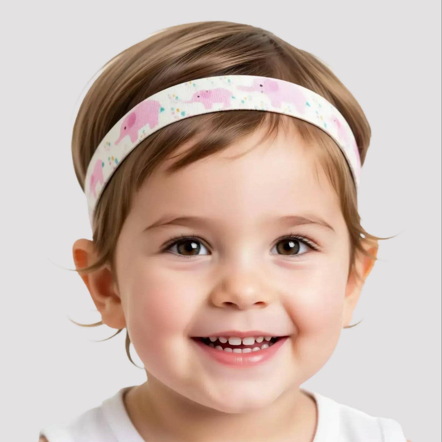 A smiling young child wearing a white hearing aid headband with a pink elephant pattern, photographed against a light gray studio background.