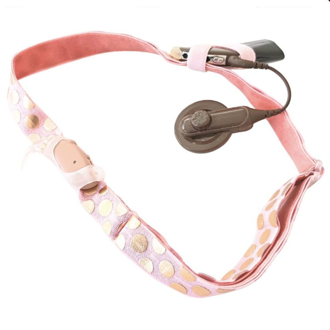 Pink headband with gold dots, with a cochlear implant and hearing aid attached