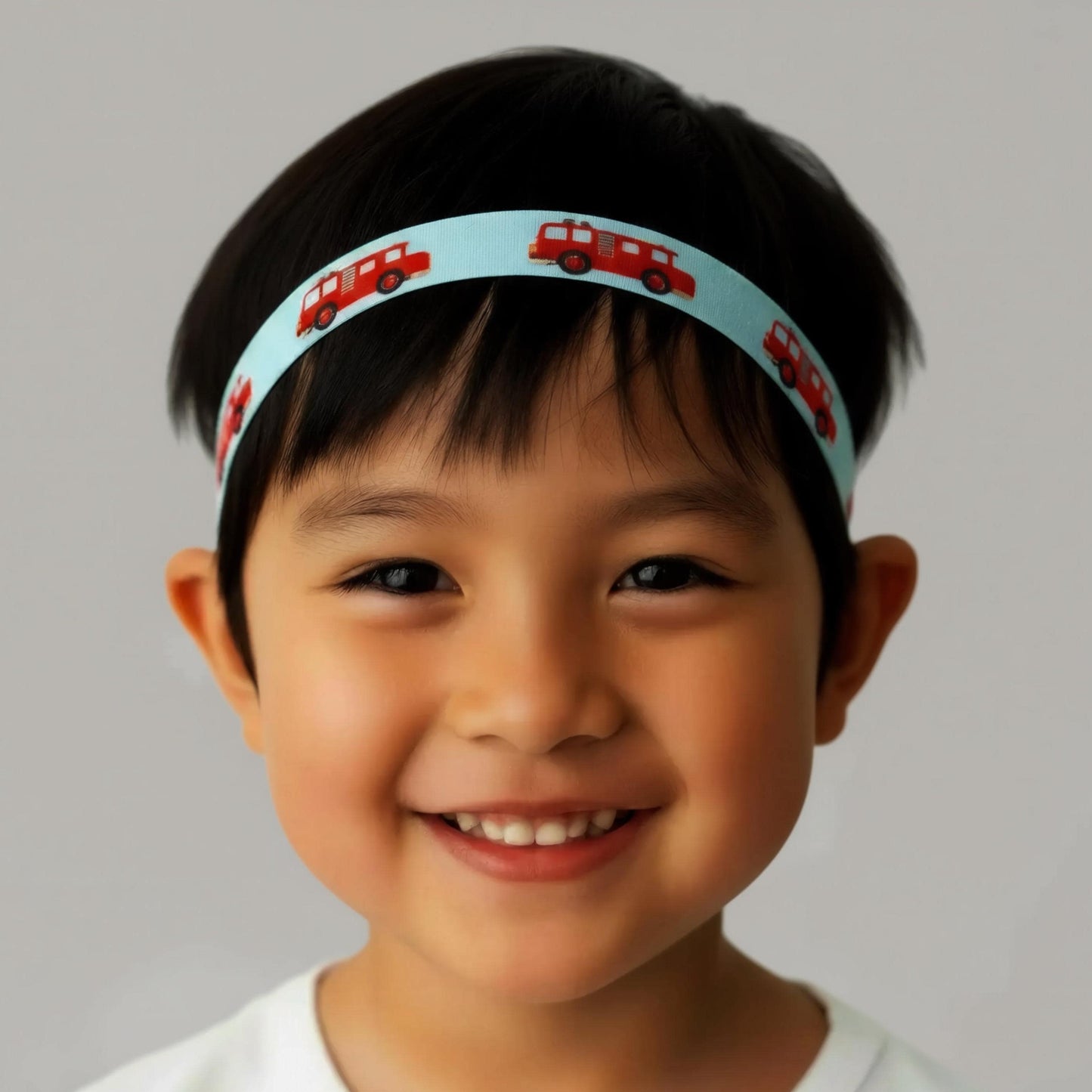 A close-up portrait of a smiling young boy wearing a light blue hearing aid cochlear implant headband printed with small red fire trucks