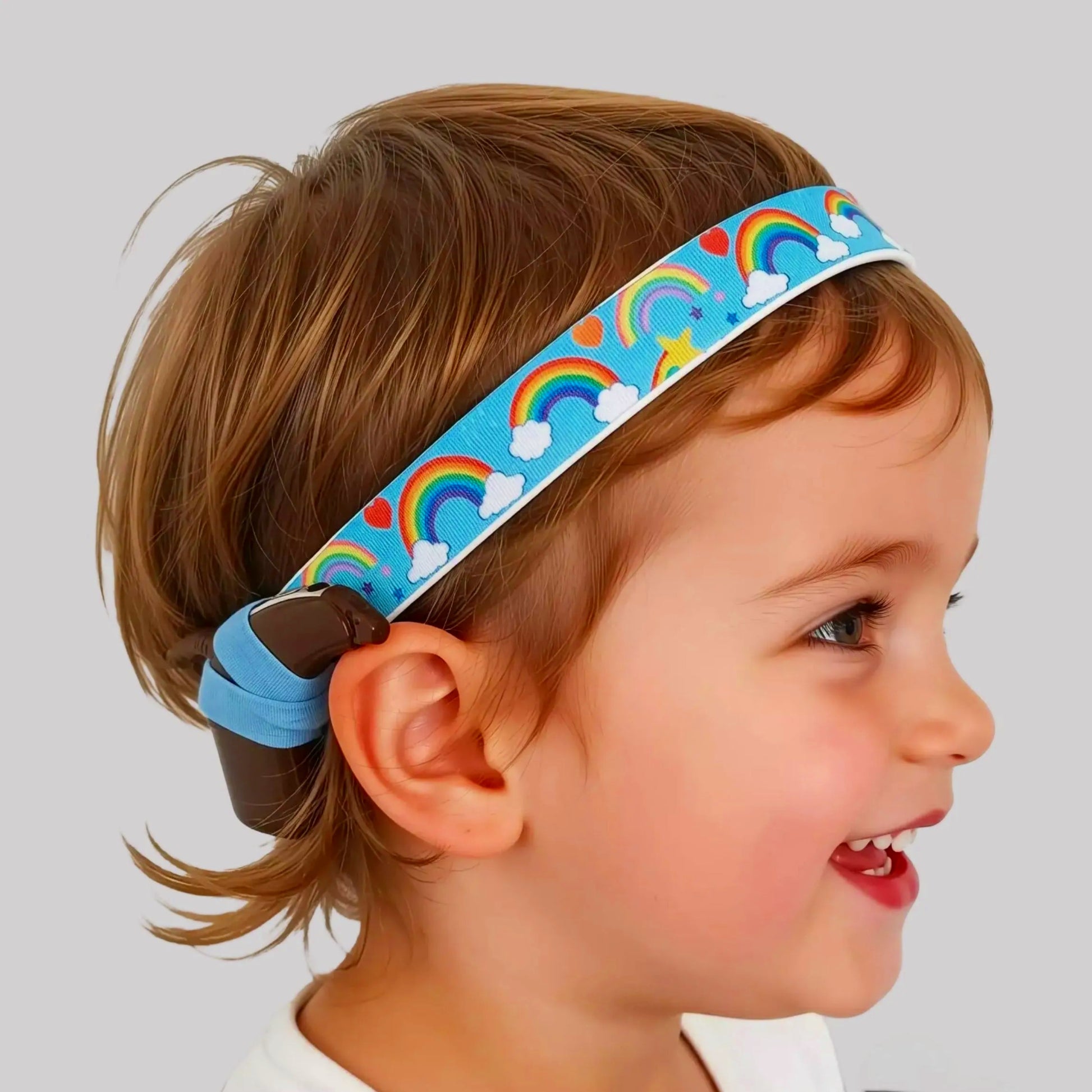 A young child wearing a light blue fabric cochlear implant hearing aid headband with a colourful rainbow and cloud pattern, shown in profile against a plain light gray background.