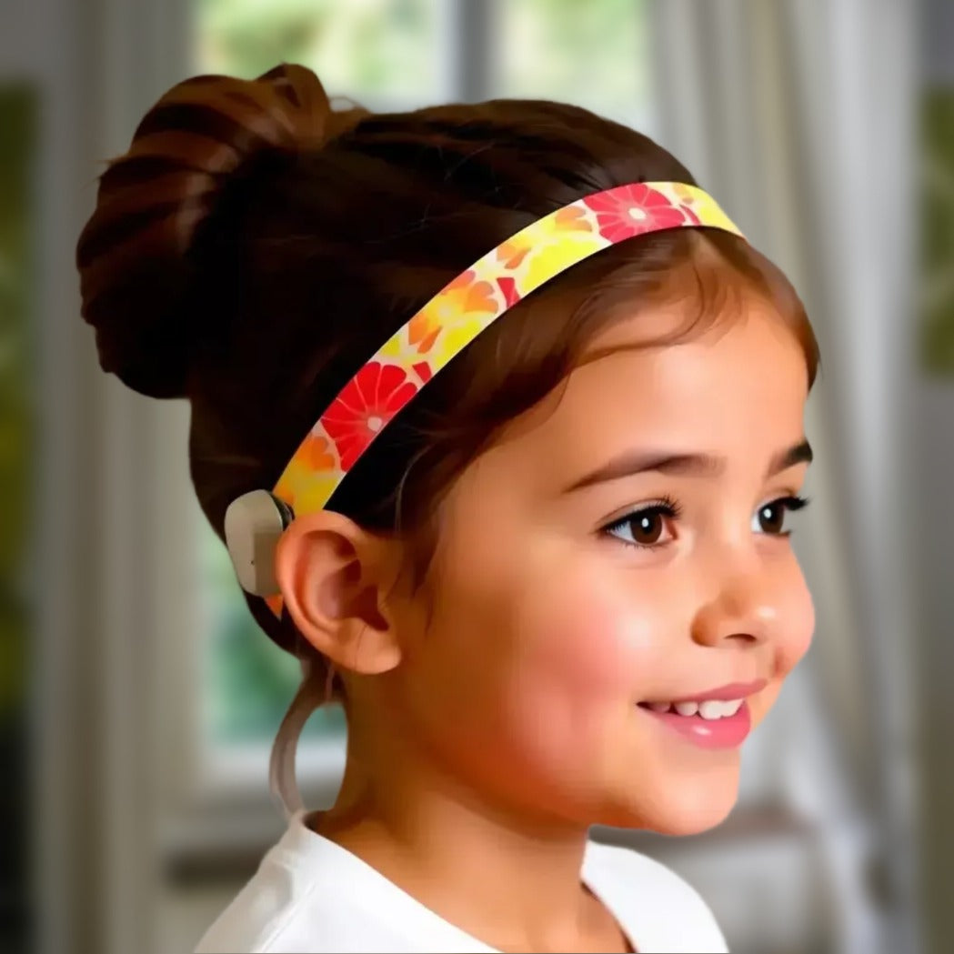 A young girl with brown hair in a bun wears a wide fabric hearing headband featuring a red, yellow, and orange citrus slice pattern.