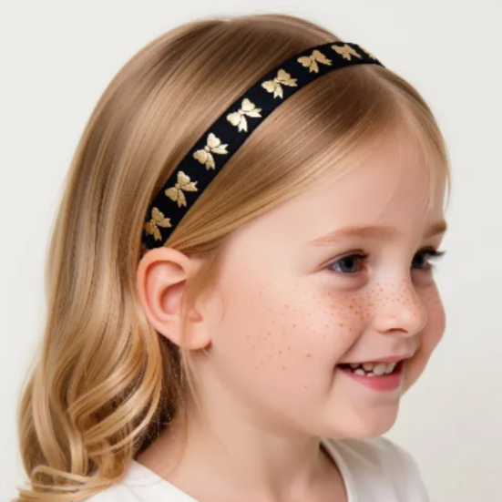 A young girl with blonde hair wearing a black hearing aid headband with gold bow designs is smiling at the camera.