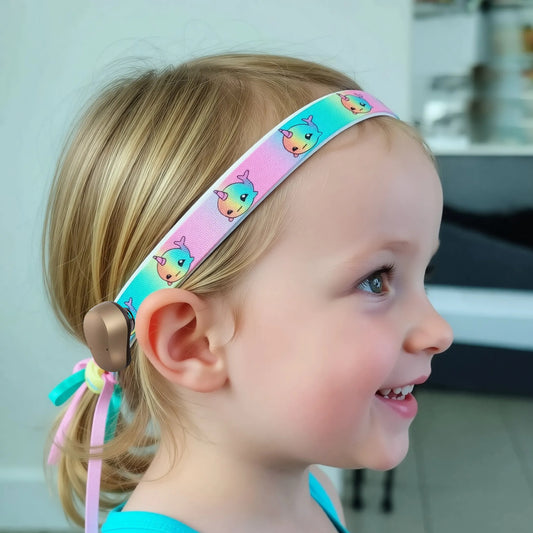 A young child in profile wearing a pastel pink and turquoise hearing aid headband with a repeating cartoon narwhal pattern