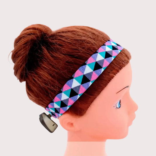 A multicolor geometric patterned ponto baha hearing aid softband headband with pink, blue, black, and white triangles, shown on a doll mannequin with brown hair in a bun against a light gray background.