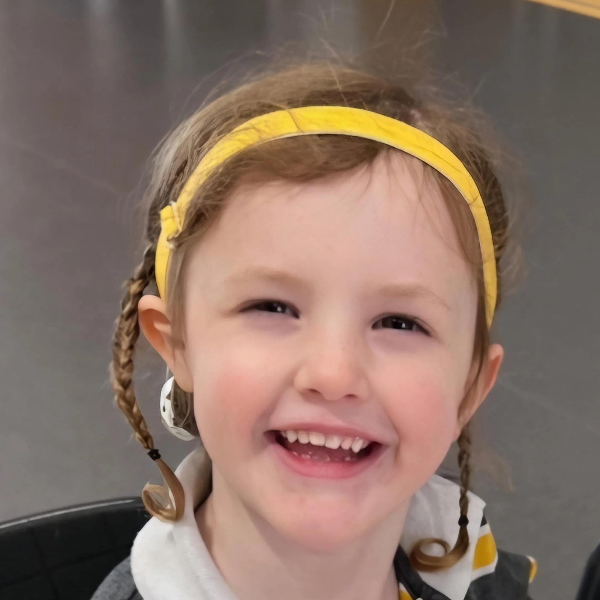 A young child with braided hair wearing a yellow fabric hearing aid headband and matching yellow-and-white clothing, shown in a close-up portrait.