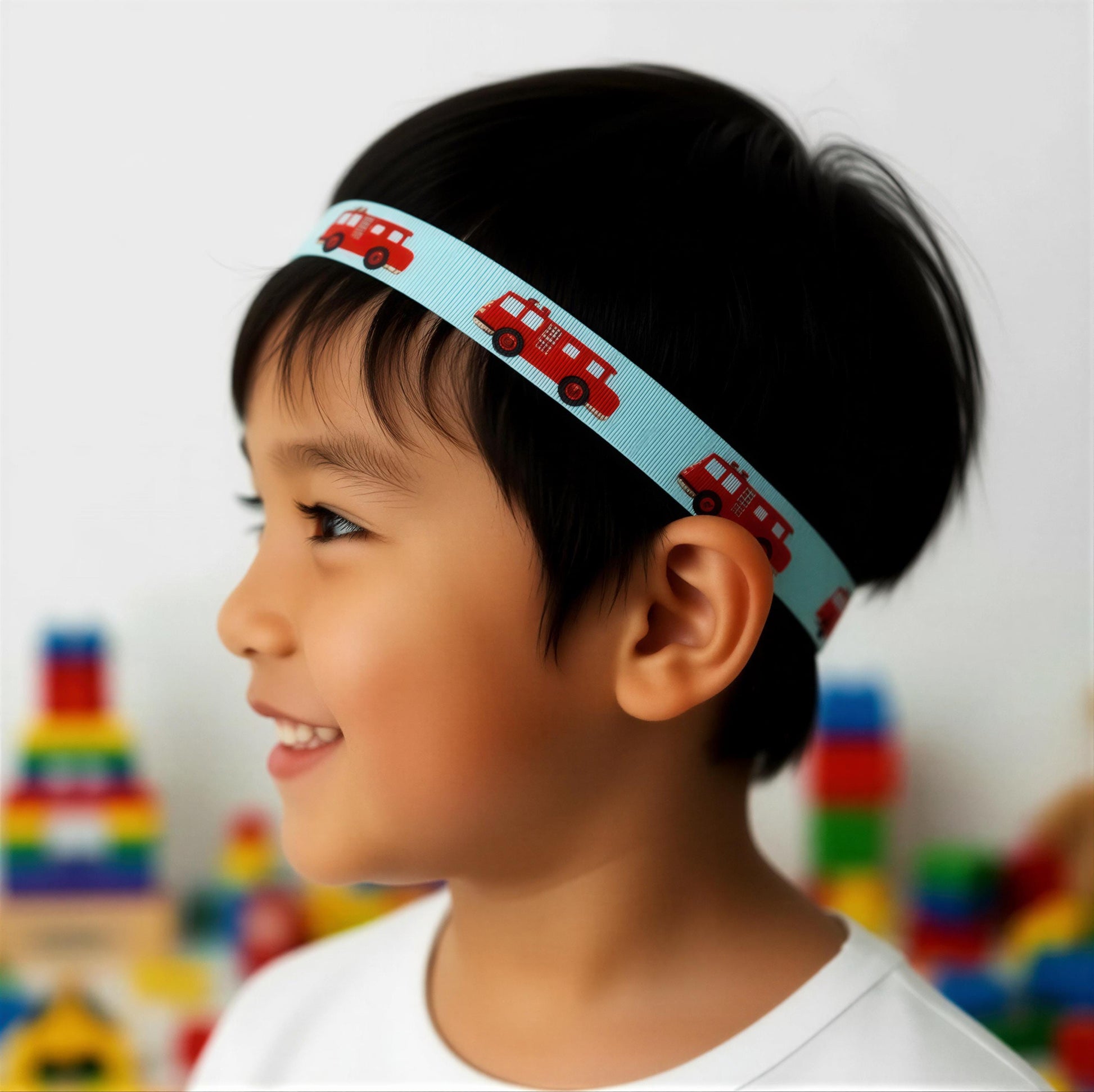 A young child in profile wearing a light blue hearing headband with a repeating pattern of red fire trucks, set against a softly blurred background of colorful toys.