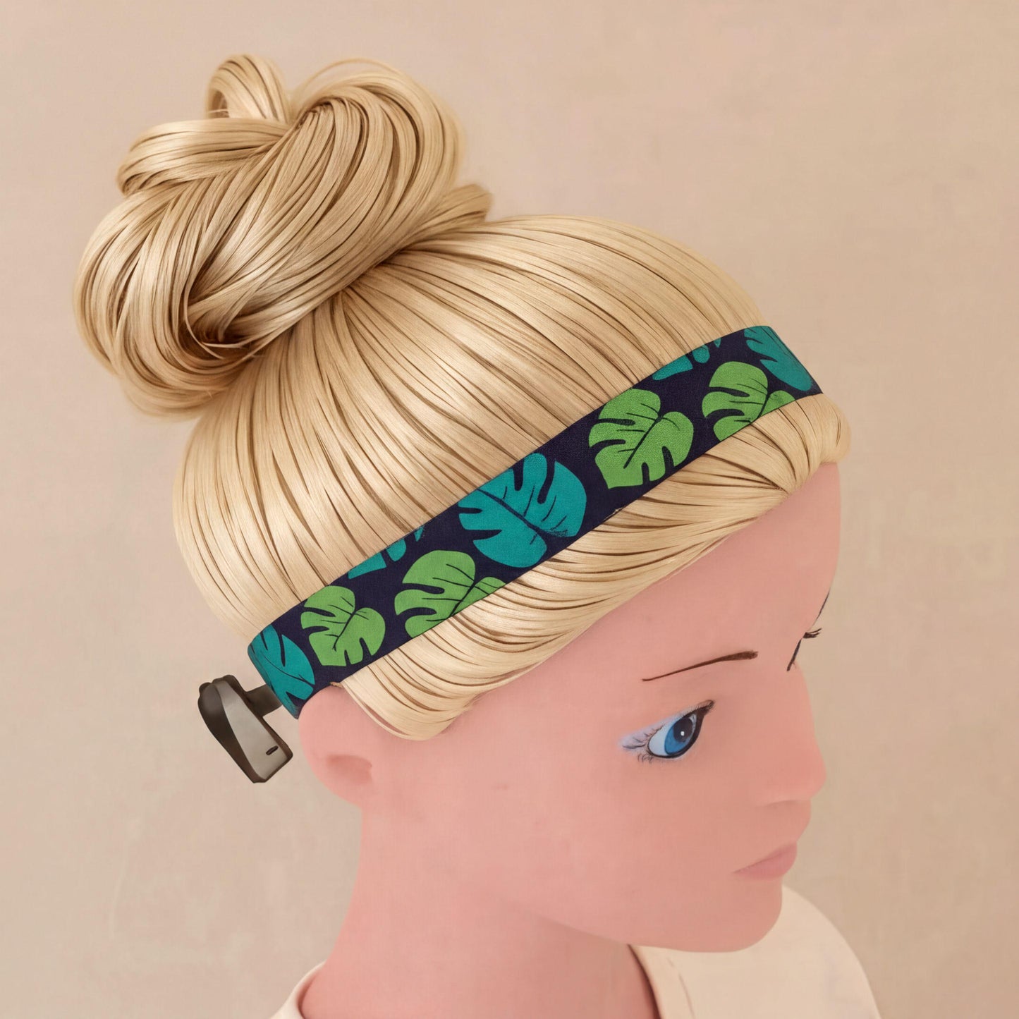 A blonde mannequin head wearing a patterned cochlear implant headband with a black base and green tropical leaf print, shown against a plain beige background.