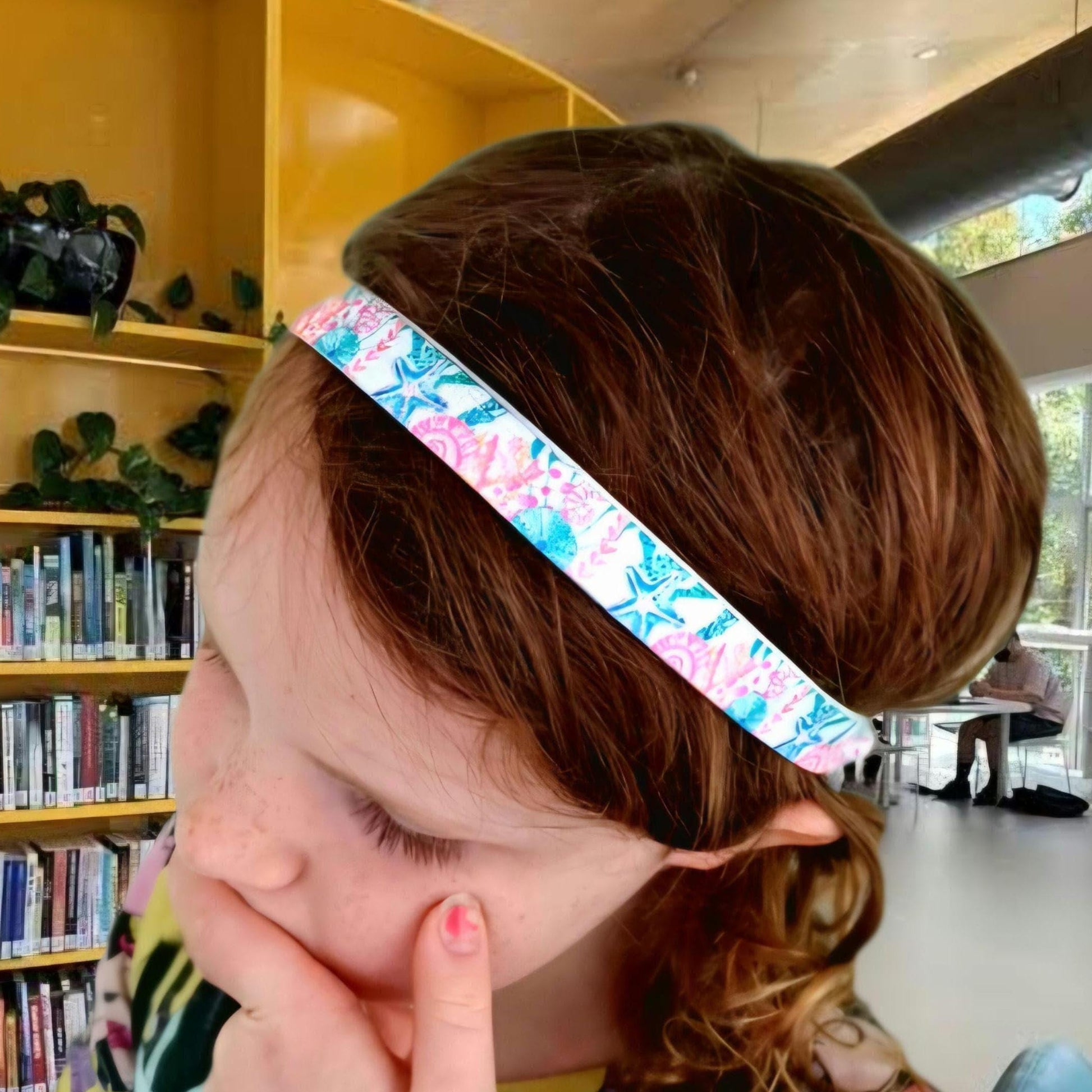 A close-up lifestyle shot of a young girl with brown hair wearing a hearing aid headband with a colourful pink, blue, and green floral pattern.