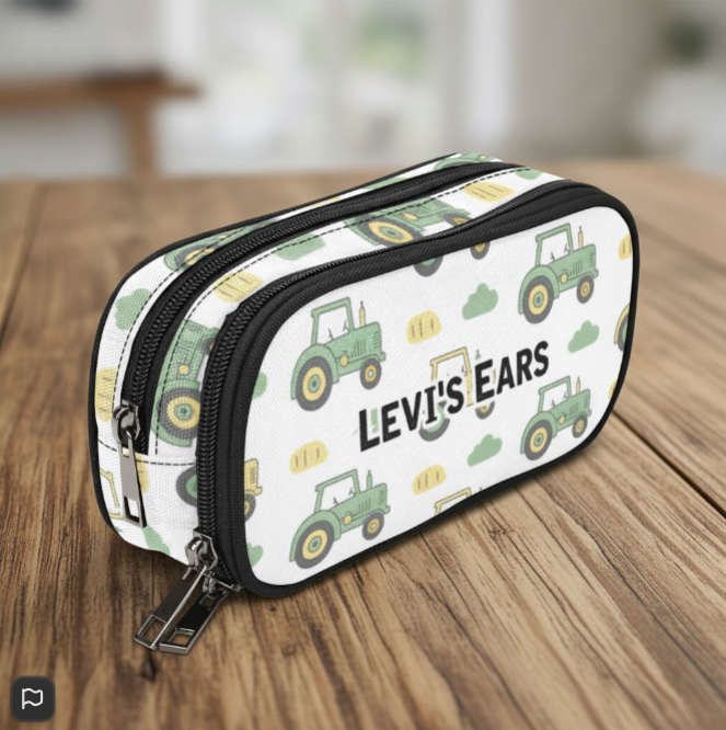 A white double-zipper storage case for cochlear implant hearing aids BAHA devices with a green tractor pattern and the text 'LEVI'S EARS' printed on the front, resting on a wooden table.