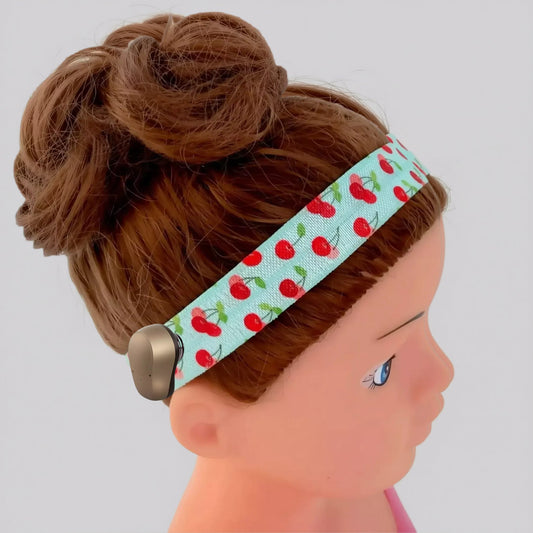 A close-up of a doll or mannequin head with brown hair in a bun, wearing a light green ponto baha hearing aid headband printed with red cherries and green stems.